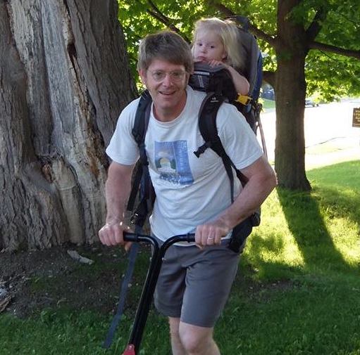 Dr. Paul pushing a lawnmower with his baby granddaughter in a carrier on his back.